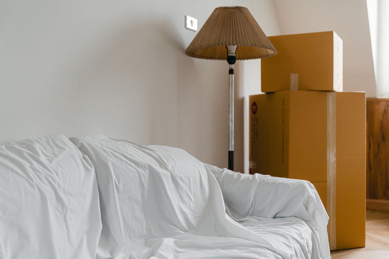 gallery-01 Covered with sheet sofa and floor lamp and packed cardboard boxes in light spacious room of new contemporary house